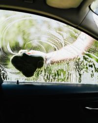Driver's side car window being washed with a soapy sponge outdoors, capturing the cleaning process.