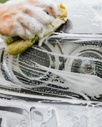 Hand scrubbing a car headlight with sponge and soap suds, detailing process.