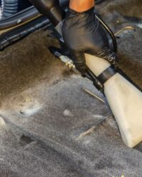 A hand in a glove using a vacuum to clean a car interior carpet, removing dirt effectively.